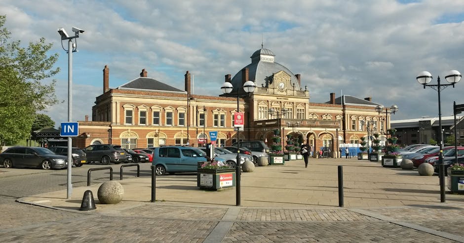 A wide view of Finsbury Park Railway Station in daylight, featuring a historic brick building with decorative architectural details, large arched windows, and a central clock. The station is surrounded by a spacious, clean paved area with black metal bollards, flower planters, and modern street lamps. Several cars are parked along the perimeter, and a few pedestrians are walking nearby. The sky overhead is partly cloudy, providing natural lighting that highlights the station's well-maintained facade. The scene reflects a tidy, inviting environment typical of a well-kept public transport hub, aligning with professional cleaning and maintenance standards highlighted by Finsbury Park Carpet Cleaning.