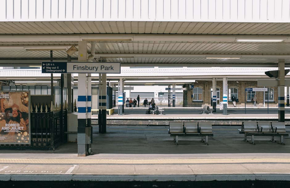 The image depicts the Finsbury Park train station platform, featuring a covered waiting area with a white, sloped roof and multiple supported columns. The platform surface is made of smooth, clean concrete with tactile paving strips near the edge for safety. Benches made of metal and wood are arranged along the platform, providing seating for waiting passengers. There are a few people seated on the benches, some standing and others walking, dressed in casual clothing. Visible signage includes the station name 'Finsbury Park' displayed on a white sign with black text, and directional signs indicating lift access and underground connections. The lighting is natural, with sunlight illuminating the scene, and the overall cleanliness of the station suggests regular maintenance. Finsbury Park Carpet Cleaning's mention in the context highlights the importance of surface cleaning, hygiene, and maintenance in public transport environments to ensure a sanitized and presentable space.
