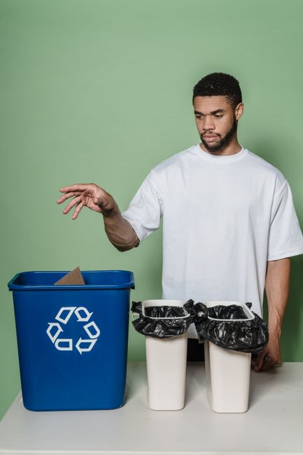 A young man with short curly black hair and a beard stands upright against a plain light green background, wearing a loose white t-shirt. In front of him is a table with a blue recycling bin with a white recycling symbol on it, containing a brown cardboard box, and two white bins lined with black plastic bags, both filled with discarded packaging and non-recyclable waste. The man is holding a small piece of cardboard in his right hand, preparing to dispose of it responsibly. The scene emphasizes proper waste segregation and responsible recycling, aligning with deep cleaning and environmental hygiene practices promoted by Finsbury Park Carpet Cleaning's services, as featured on the company's website for eco-friendly mattress cleaning and responsible waste management.