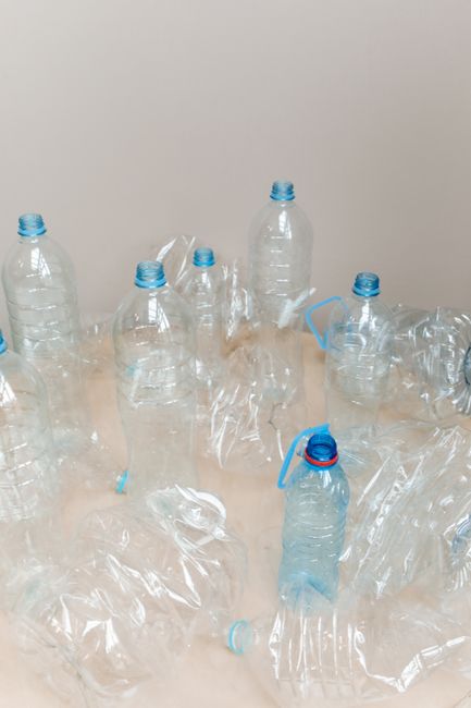 A collection of clear plastic bottles with blue caps and handles placed on a light-colored tabletop, some of which are crushed or deformed, indicating they have been emptied. A few plastic wrappers or bags are also visible among the bottles. The background is a plain neutral wall with soft, natural lighting, reflecting a clean and organized environment. The scene depicts a responsible recycling process as part of eco-friendly cleaning practices, aligning with Finsbury Park Carpet Cleaning's commitment to environmental sustainability and surface cleaning or waste management in domestic or commercial settings.