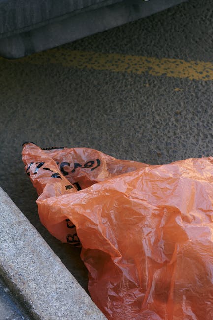 An orange plastic waste bag resting on an asphalt surface at the edge of a sidewalk, with a concrete curb and a grey fabric-covered bench or structure in the background. The ground shows patches of dust and dirt, with a yellow painted line visible on the road. The image emphasizes waste disposal in an outdoor urban setting, relevant to maintaining cleanliness standards in accordance with Haringey Council rules as highlighted by Finsbury Park Carpet Cleaning's cleaning services. The scene features natural lighting with no visible cleaning tools or products present, focusing on the importance of proper waste management for hygiene and environmental responsibility.