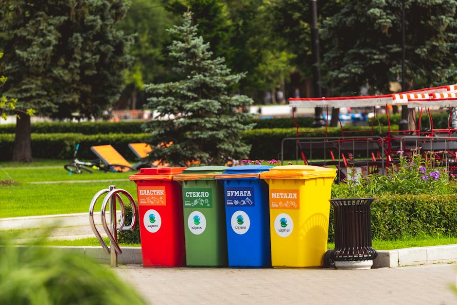 A group of four wheeled waste bins in blue, red, yellow, and black, positioned on a snow-covered ground in a wooded outdoor area with tall trees. The scene appears cold and wintry, with snow blanketing the grass and branches, and trees with some snow on their trunks and branches. The background shows dense trees with a mixture of snow and sparse foliage, under cloudy skies. The bins are arranged in a row near a large tree, and there is minimal human activity visible. This image emphasizes outdoor waste disposal in a natural setting, aligning with the importance of following local waste management rules, as discussed on finsburyparkcarpetcleaning.com’s page about Haringey Council regulations for cleaning waste and disposal in N4, FINSBURY PARK. Finsbury Park Carpet Cleaning promotes proper hygiene and waste practices as part of comprehensive cleaning and sanitisation services.