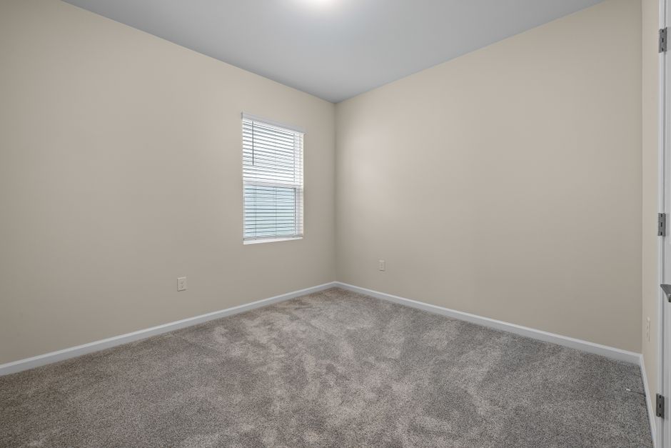 Empty room with beige painted walls and a light gray carpeted floor, illuminated by natural light coming through a double-pane window with white blinds. The room features white baseboards along the perimeter and a closed white door on the right side. The smooth ceiling is painted in a light color. The space appears clean and well-maintained, suitable for surface cleaning or domestic deep cleaning, with no visible dust or clutter. This image exemplifies a freshly cleaned, vacant room prepared for occupied use, as might be treated by Finsbury Park Carpet Cleaning in accordance with Islington Council permissions for listed homes.