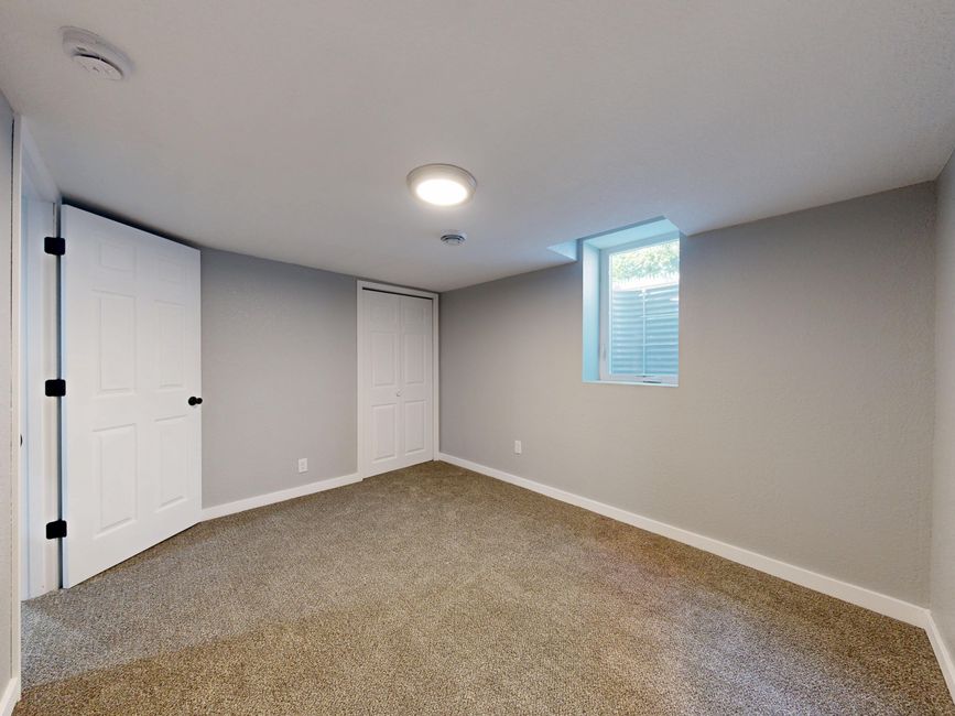A clean, empty bedroom with light gray painted walls and a beige carpeted floor. The room features a small rectangular window with white blinds, allowing natural light to brighten the space. There are two white doors: one with a black handle on the left, possibly leading to a closet or another room, and a closet with a sliding door on the adjacent wall. A circular ceiling light provides additional illumination, and a white electrical socket is visible near the baseboard. The overall appearance suggests recent surface cleaning and maintenance, typical of professional domestic cleaning services provided by Finsbury Park Carpet Cleaning, aligning with their expertise in surface cleaning and hygiene for residential spaces.