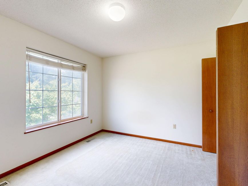 Empty room with beige painted walls and a light gray carpeted floor, illuminated by natural light coming through a double-pane window with white blinds. The room features white baseboards along the perimeter and a closed white door on the right side. The smooth ceiling is painted in a light color. The space appears clean and well-maintained, suitable for surface cleaning or domestic deep cleaning, with no visible dust or clutter. This image exemplifies a freshly cleaned, vacant room prepared for occupied use, as might be treated by Finsbury Park Carpet Cleaning in accordance with Islington Council permissions for listed homes.