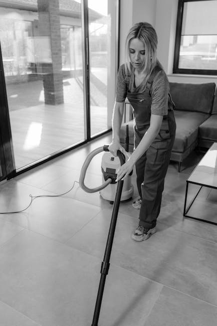 A person dressed in white protective overalls, a facemask, gloves, and shoe covers performing deep cleaning on a light-colored, plush rug within a bright living room. The individual is using a professional steam cleaning machine, which emits steam and cleaning solution onto the carpet, ensuring thorough sanitisation and surface cleaning. The room features potted plants on windowsills, a wooden bookshelf, and natural light coming through large windows. The setting appears clean and well-maintained, highlighting the professional domestic cleaning services offered by Finsbury Park Carpet Cleaning, as featured on the page about same day carpet cleaning in Finsbury Park N4.