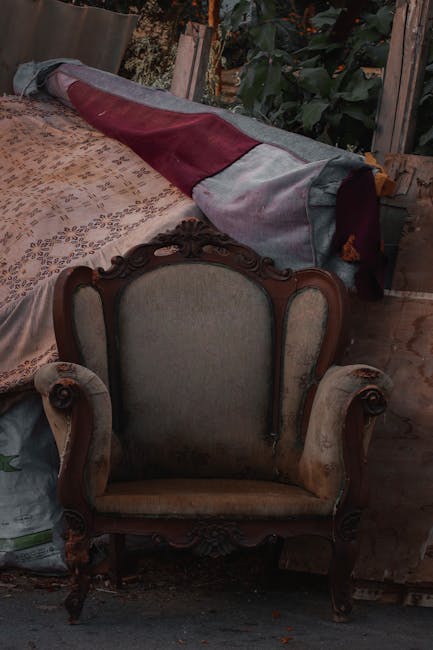 A vintage armchair with ornate wooden carvings and upholstered in light beige fabric, positioned in front of a cluttered space. Behind the chair, there is a table covered with a patterned cloth, and a partially visible piece of furniture with a red and grey fabric draped over it. The background includes dark foliage and wooden fencing, indicating an outdoor or semi-outdoor setting. The lighting is natural but slightly subdued, highlighting the intricate details of the armchair's woodwork and the surrounding objects, suggesting a scene of domestic tidying or preparation for cleaning, consistent with services provided by Finsbury Park Carpet Cleaning on Stroud Green Road. This setting exemplifies the importance of surface cleaning and maintenance to preserve furniture and household items.