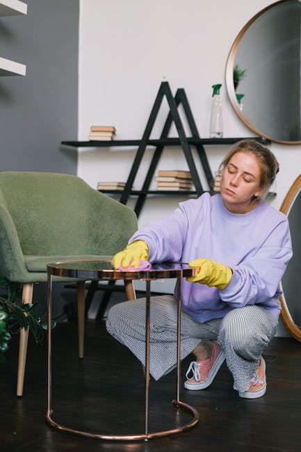 A woman with long, light brown hair tied in a bun is leaning over a sofa armrest, engaged in domestic cleaning within a living room. She is wearing a greyish-green casual jacket with rolled-up sleeves. The sofa has dark green fabric upholstery, and positioned on it are red and grey cushions. Behind her, a white brick wall features a large mirror, and a bookshelf with colorful books is visible. The room is brightly lit by natural light and a ceiling light fixture, creating a warm, inviting atmosphere. This scene illustrates a surface cleaning or upholstery cleaning activity, exemplifying professional deep cleaning services provided by Finsbury Park Carpet Cleaning, as highlighted on the Stroud Green Road upholstery cleaning experts page.